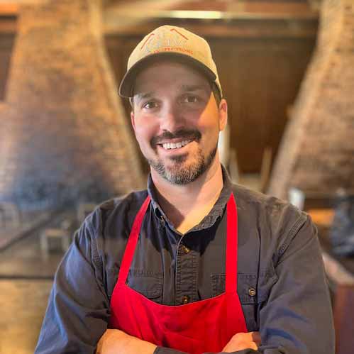 Portrait of Luke Askew, owner of Holcombs Bar-B-Que wearing a red chef's apron and smiling at the camera.