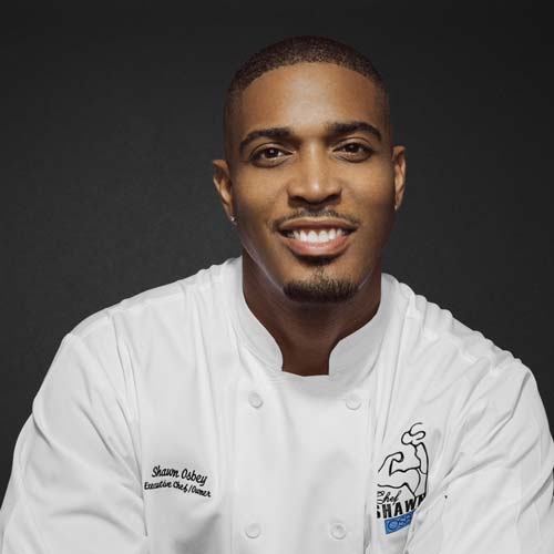 Chef Shawn Osbey headshot in chef's jacket against a dark grey background.