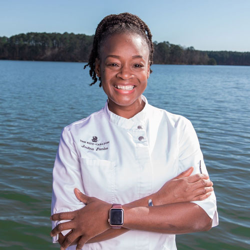 Sous Chef Jestina Pambu smiling in her white chef's coat in front of Lake Oconee.