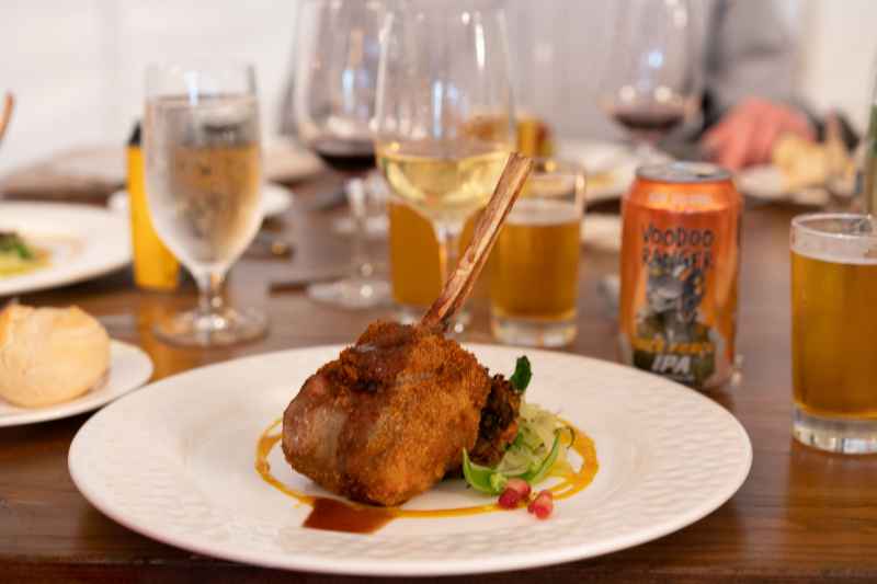 A succulent lamb chop sits on a beautifully presented dish on table with craft beer glasses at the Hops & Vine Dinner.